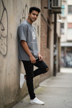 A man wearing a gray graphic t-shirt, black ripped jeans, and white sneakers leans against a concrete wall in an urban alleyway.