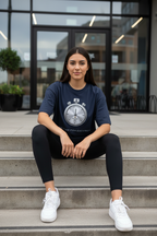 A woman sits on concrete steps in front of a modern glass building, wearing a navy blue graphic t-shirt with a white stopwatch design and the text 'TIME FOR EXCUSE', black leggings, and white sneakers.