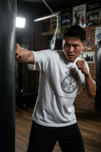 A man in a boxing gym wearing a white graphic t-shirt and black pants, punching a heavy bag. The t-shirt features a circular stopwatch graphic with the text 'NO TIME FOR EXCUSE'.