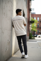 A man wearing a cream-colored t-shirt with 'PROGRESS DELIVERED DAILY' printed in black on the back, paired with dark gray pants and white sneakers, leaning against a concrete wall on an urban sidewalk.