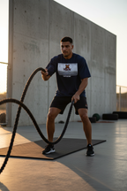 A man in a navy blue athletic t-shirt and black shorts is performing battle rope exercises on a concrete outdoor gym floor. The t-shirt features a white rectangular graphic on the chest with a red logo and text.
