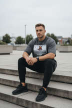 A man sitting on outdoor concrete steps wearing a dark gray graphic t-shirt, black sweatpants, and black sneakers.