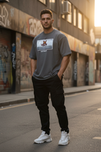 A man standing on an urban street wearing a gray graphic t-shirt, black cargo pants, and white sneakers. The background features graffiti-covered building shutters and warm golden-hour sunlight.