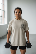 Woman holding dumbbells wearing a beige t-shirt with a logo in a room with large windows.