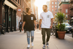 A man and a woman walking side-by-side on a city sidewalk during golden hour, wearing matching graphic t-shirts from the brand From Me Fitness Apparel. The woman wears a black t-shirt and distressed light-wash jeans, while the man wears a white t-shirt and dark olive pants.