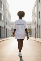 A person viewed from behind walking down a sunlit urban street, wearing an oversized white T-shirt dress with small black text across the upper back.